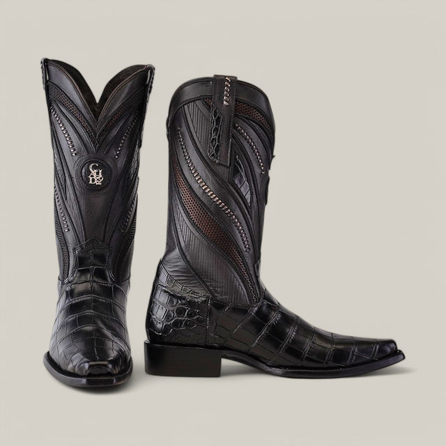 The IMPERIO Alligator Belly Black Boots feature a glossy alligator texture, intricate stitching, decorative zippers on a pale backdrop, and a narrow square toe. One boot stands upright while the other is slightly angled to emphasize its elegance.