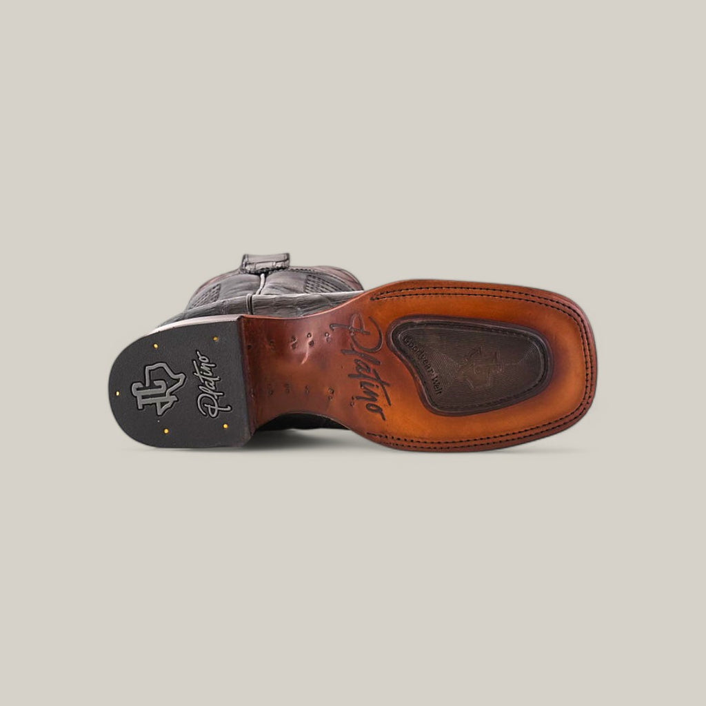 The image displays the underside of a Exotic American Alligator - Dark Gray - Square Toe boot laying on its side. The brown sole features distinctive stitching and an embossed logo on the heel, while the textured black heel has two small gold studs, showcasing its intricate design.