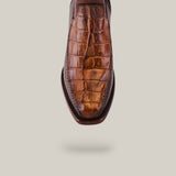 A brown shoe with crocodile skin texture is displayed against a plain background. The Exotic American Alligator Tail - Fawn boot, featuring a handcrafted design, faces the viewer toe-first to emphasize its detailed pattern and stitching.