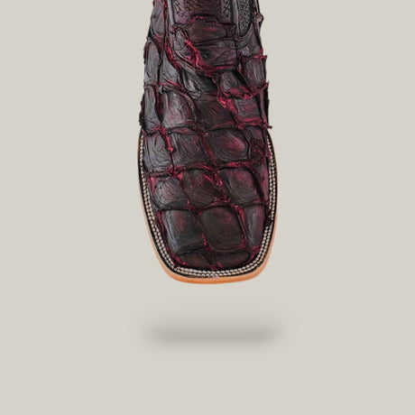 Close-up of a cowboy boot with a textured matte black cherry Pirarucu fish pattern and a square-toe design. Subtle stitching accents the edges, while the plain light gray background complements this exotic leather masterpiece.