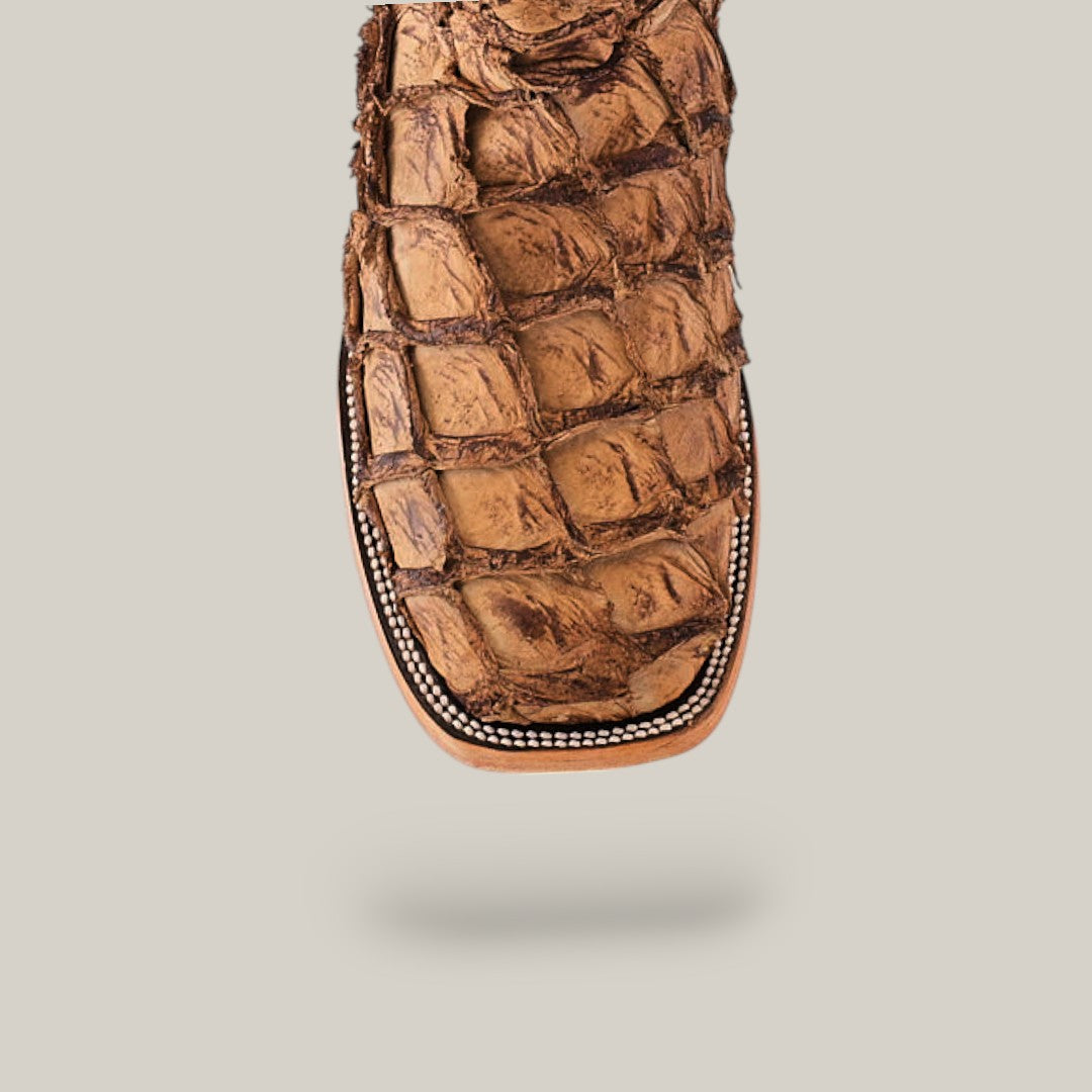 A close-up of the Exotic Pirarucu Fish - Matte Orix - Square Toe boot showcases its tan, rugged texture reminiscent of Amazonian Pirarucu skin, with a square toe and decorative stitching. The neutral background accentuates its unique craftsmanship.