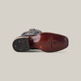 The image displays the sole of the Exotic Ostrich - Black - Narrow Square Toe boot with a wooden finish, embodying Western heritage. A black heel with grip patterns and logos adds detail to this genuine leather masterpiece, set against a plain background.