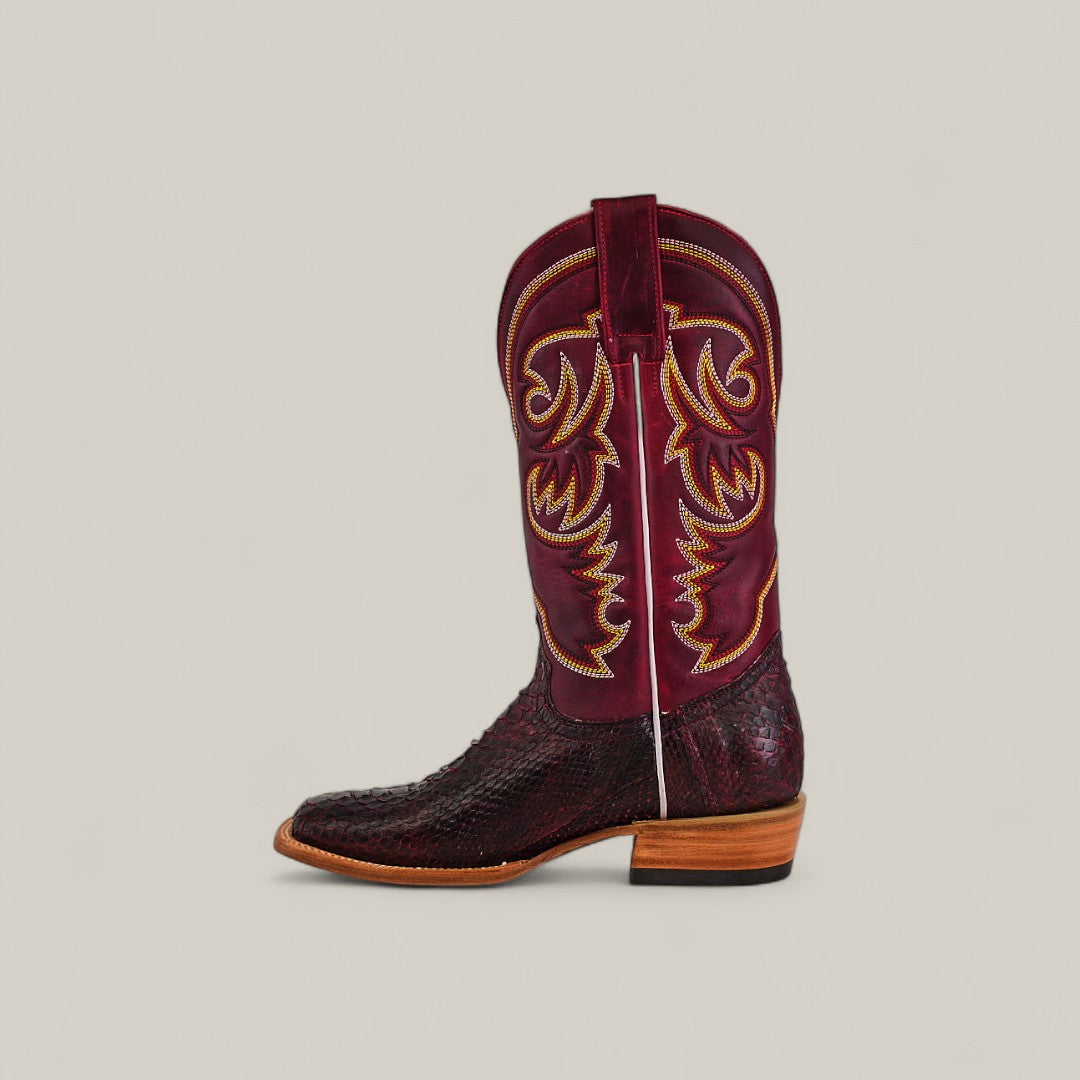 A single Exotic Python - Blackcherry - Square Toe boot, featuring intricate yellow stitching, textured finish, pull strap, and light brown stacked heel, is displayed on a plain beige background.