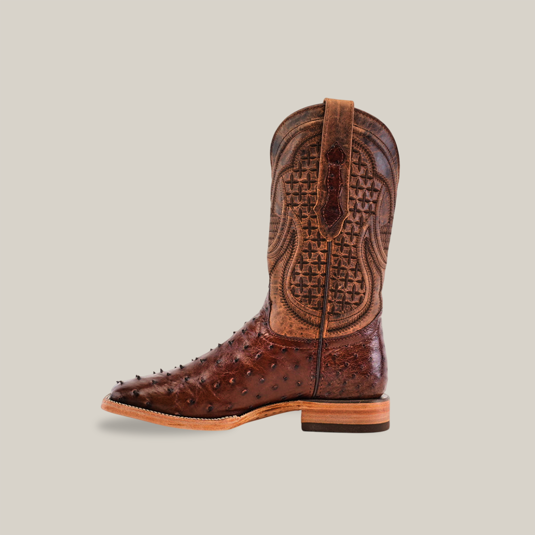 The Exotic Full Quilt Ostrich Boot - Brown - Square Toe features intricate tooling and textures in exotic ostrich leather. It has decorative shaft patterns, a smooth sole, and raised toe bumps that stand out against a plain beige backdrop.