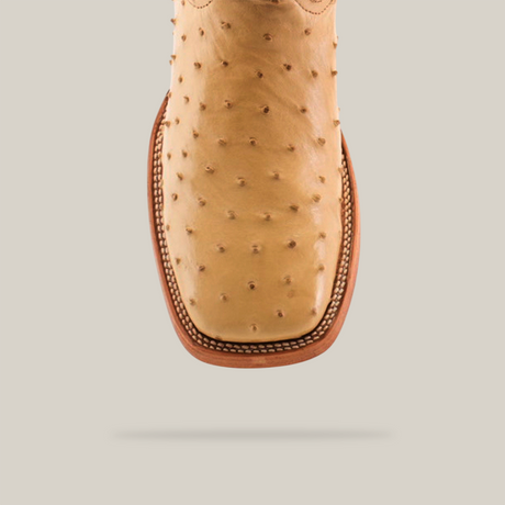 A close-up of the Exotic Full Quilt Ostrich - Orix cowboy boot highlights its tan ostrich leather with distinctive bumps, square toe design, and intricate stitching around the toe and sole, all set against a plain gray background.