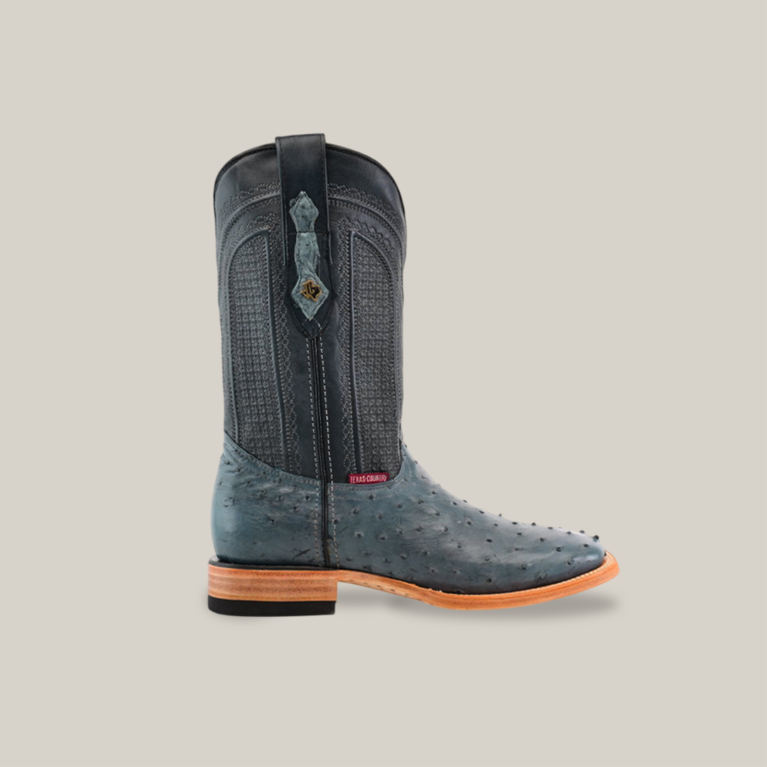 The Exotic Full Quilt Ostrich - Blue Jean - Square Toe cowboy boot showcases a dark blue ostrich leather foot like blue jeans, with a textured shaft. It features a wooden heel and sole, and the tag reads Handcrafted. The boot stands upright against a beige background.