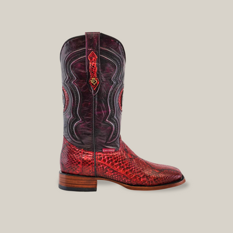 The Exotic Python - Ruby - Square Toe is a single cowboy boot featuring a ruby snakeskin texture, intricate dark shaft stitching, and wooden heel and sole accents. Set on a plain beige background, it showcases decorative patterns that blend elegance with rustic charm.