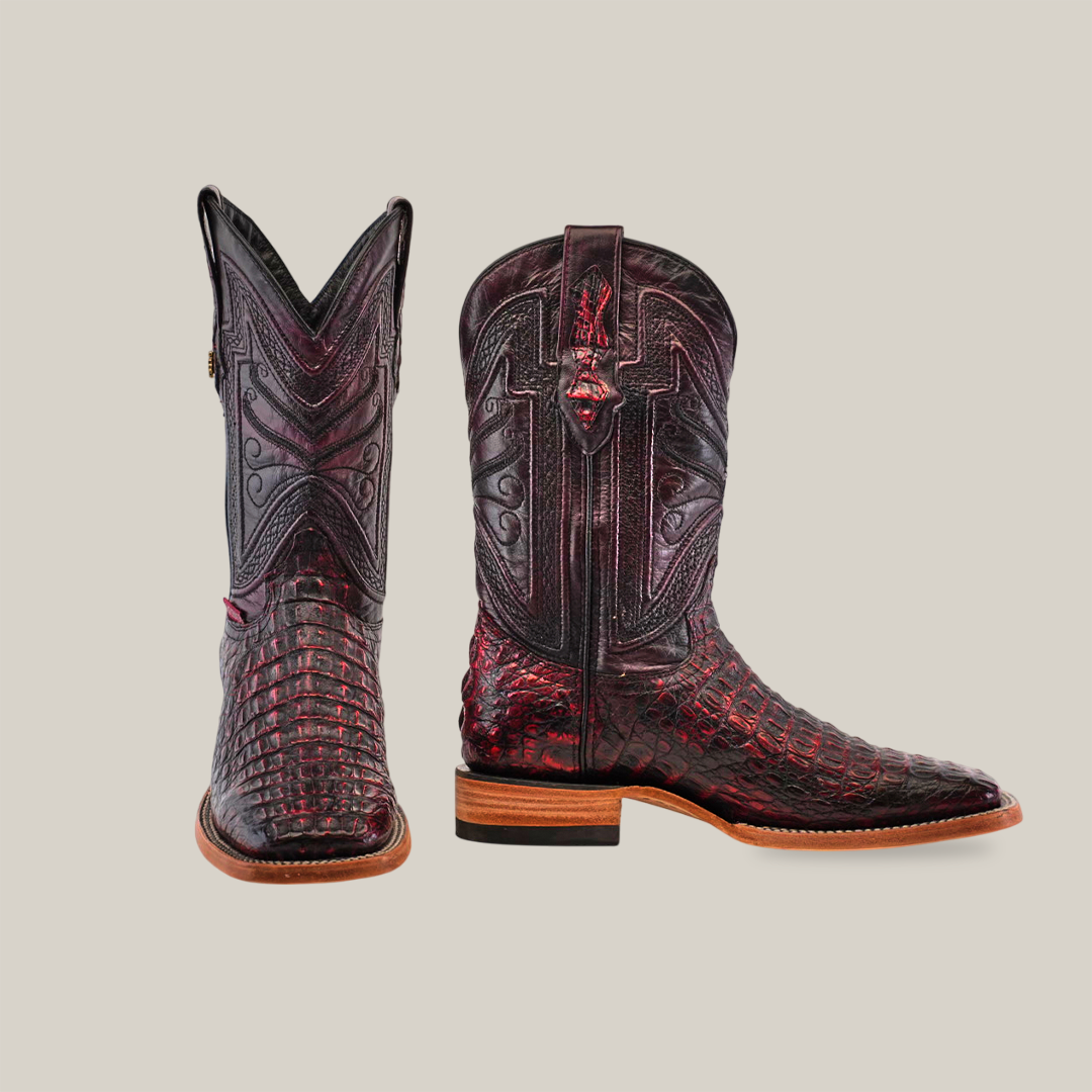 The Exotic Caiman Horn Back - Black Cherry boots feature a dark red and black crocodile texture with intricate designs. These Square Toe boots highlight detailed stitching and come with a wooden sole featuring a slightly raised heel, showcased with one boot facing forward and one to the side.