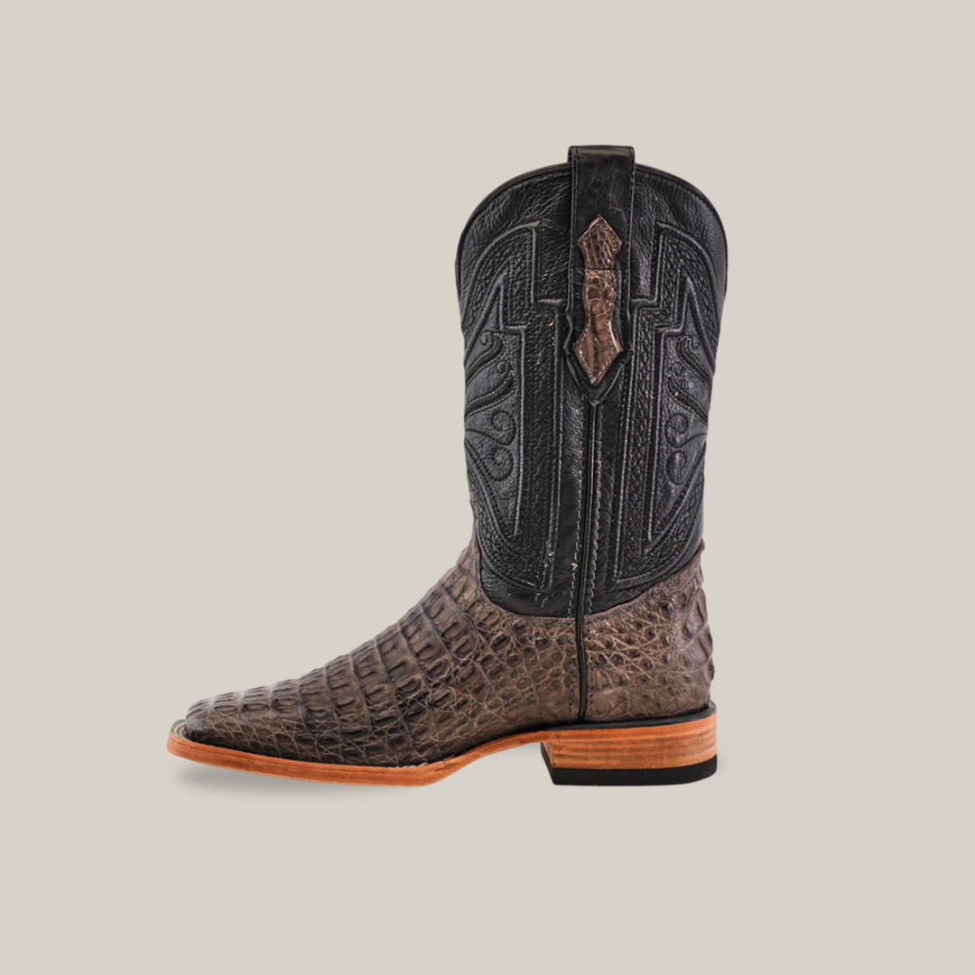 A single Exotic Caiman Horn Back - Serpentine cowboy boot has a black shaft with decorative stitching and a caiman leather textured lower part reminiscent of alligator skin. It features a light brown sole, stacked wooden heel, and square toe design against a plain background.