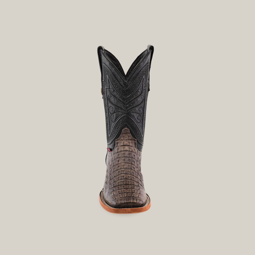 The Exotic Caiman Horn Back - Serpentine boot features a forward-facing black upper with intricate stitching, a textured brown lower made of genuine caiman leather, and a light brown sole. It is displayed against a plain beige background.