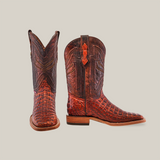 A pair of Exotic Caiman Horn Back cowboy boots in Smoke Cogñac with a square toe, featuring intricate patterns and detailed stitching. One boot stands upright while the other lies on its side, displaying a wooden sole against a plain light beige background, highlighting their classic style.