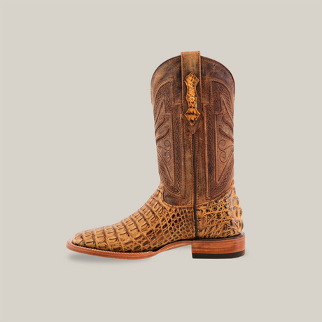 A handcrafted Exotic Caiman Horn Back - Biza Camel - Square Toe boot with intricate stitching and a textured reptile-like pattern is set against a plain beige background, featuring a pull tab at the top and a wooden heel.