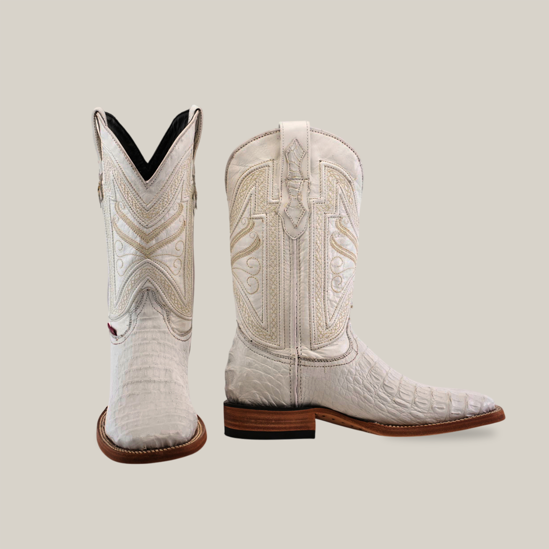 The Exotic Caiman Horn Back boots in white, crafted from premium leather, showcase detailed embroidery on a neutral background. The left boot has a square toe and dark interior, while the right boot highlights quality craftsmanship with its wooden heel and sole.