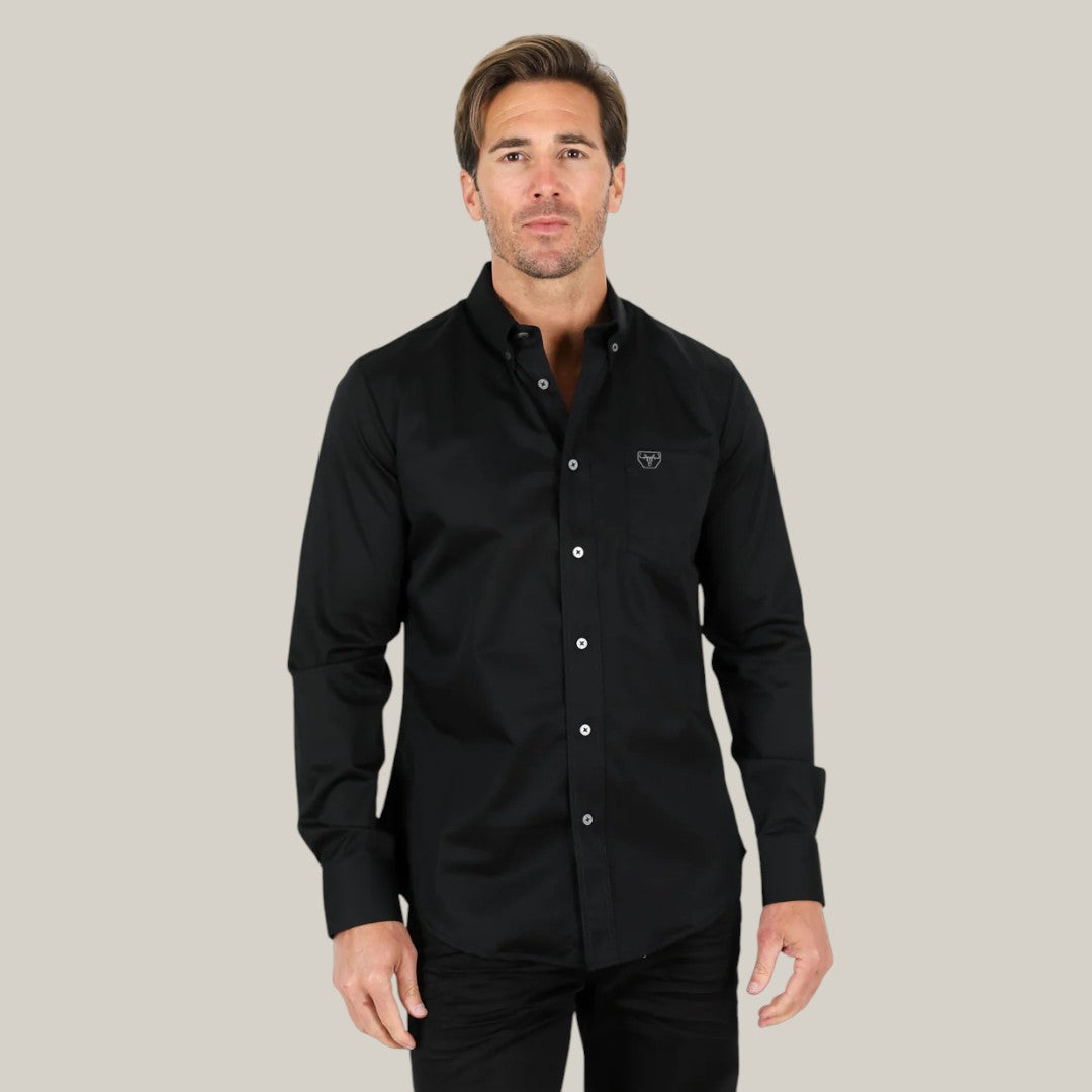 A man in a Mens Single Pocket Logo Modern Fit Stretch Dress Shirt - Black (CTL9258) and black pants stands against a light gray background. The shirts small embroidered logo, paired with his short hair and neutral expression, enhances the contemporary style.