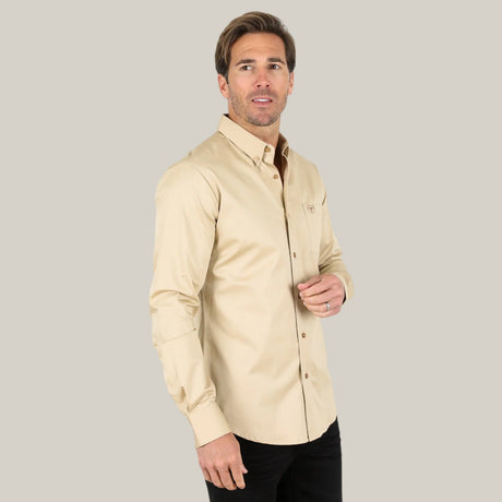 A person in a beige Men’s Single Pocket Logo Modern Fit Stretch Dress Shirt (CTL9261) and black pants stands against a light gray background, gazing to the side with a neutral expression.