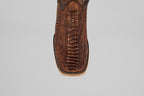 Close-up top view of the Exotic Full Quilt Ostrich Leg - Rustic Brick, a square-toed cowboy boot in brown with textured, patterned ostrich leg leather and detailed edge stitching. Set against a plain light gray background.