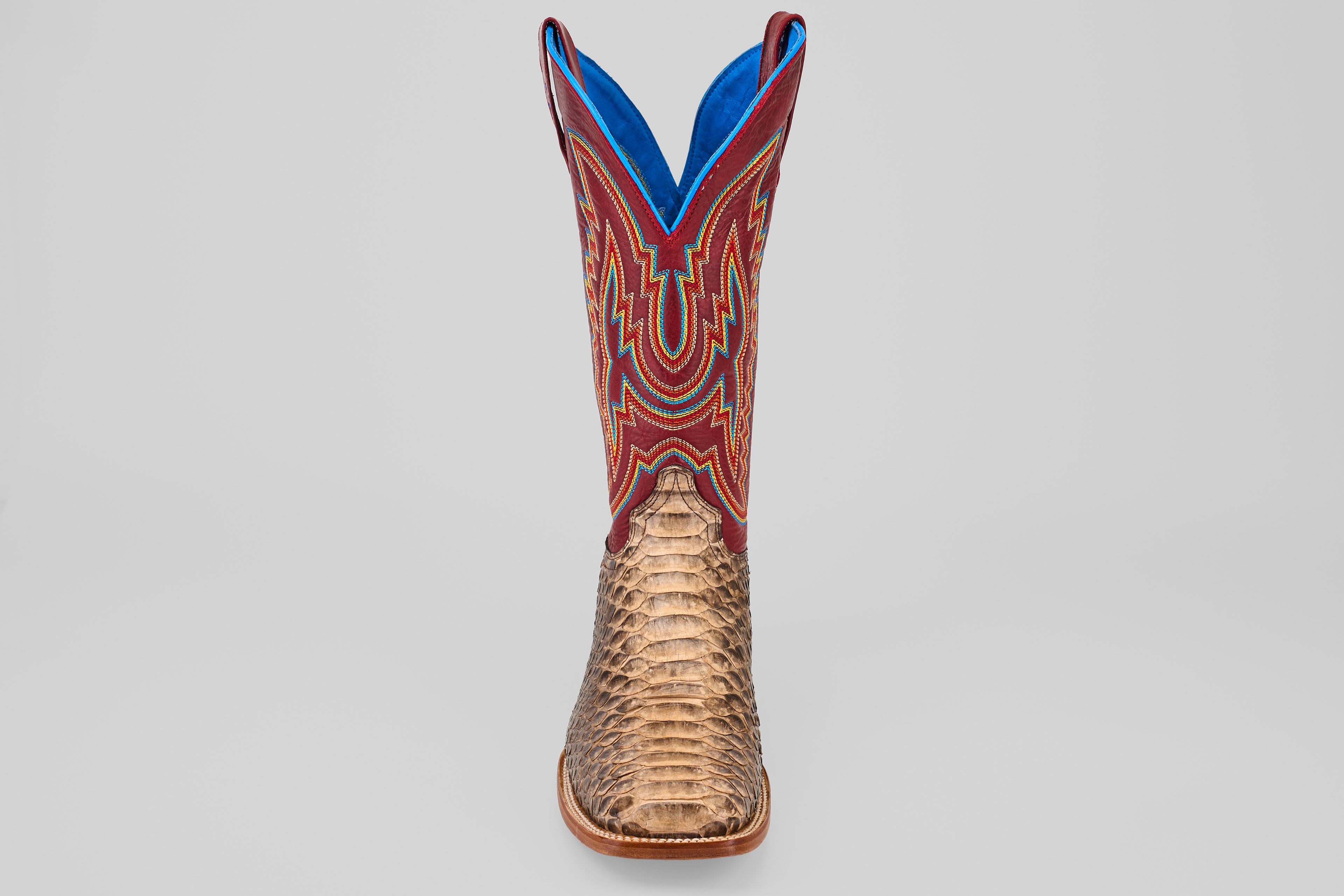 The Barlow - Taupe - Square Toe men’s statement boot features a textured taupe python leather lower, a vibrant red upper with blue and yellow stitching, and is displayed front-facing against a plain gray background.