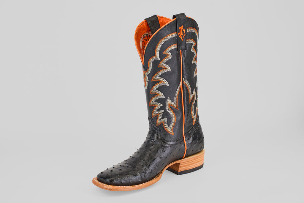 The Steele - Black - Square Toe boot features a single black ostrich leather foot, orange and white decorative stitching, an orange-trimmed pull tab, and a wooden sole with a slight heel for a touch of luxury.