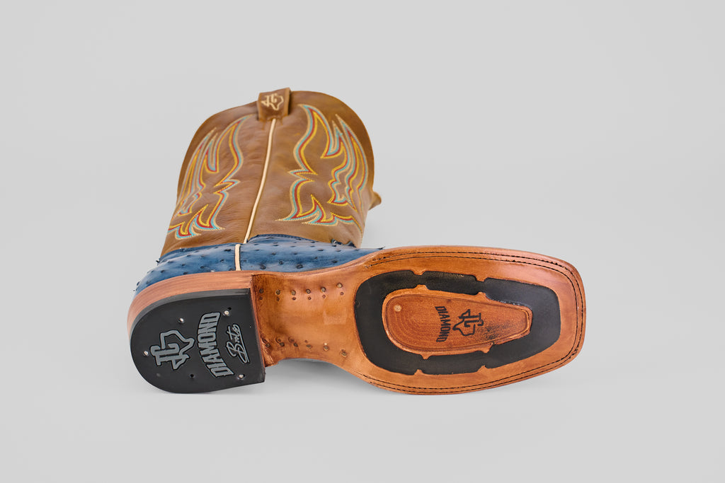 The Ridge - Blue - Square Toe boot from the Platinum Collection features a tan leather sole, blue and tan upper, yellow stitching, and a black rubber heel labeled “Diamond.” It is shown lying on its side with the sole facing up.