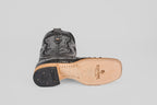 The Austin - Black - Square Toe boot lies on its side, highlighting detailed stitching and a tan leather sole with the Texas Country logo. Made from suave negro avestruz, it features a black heel adorned with small brass nails.