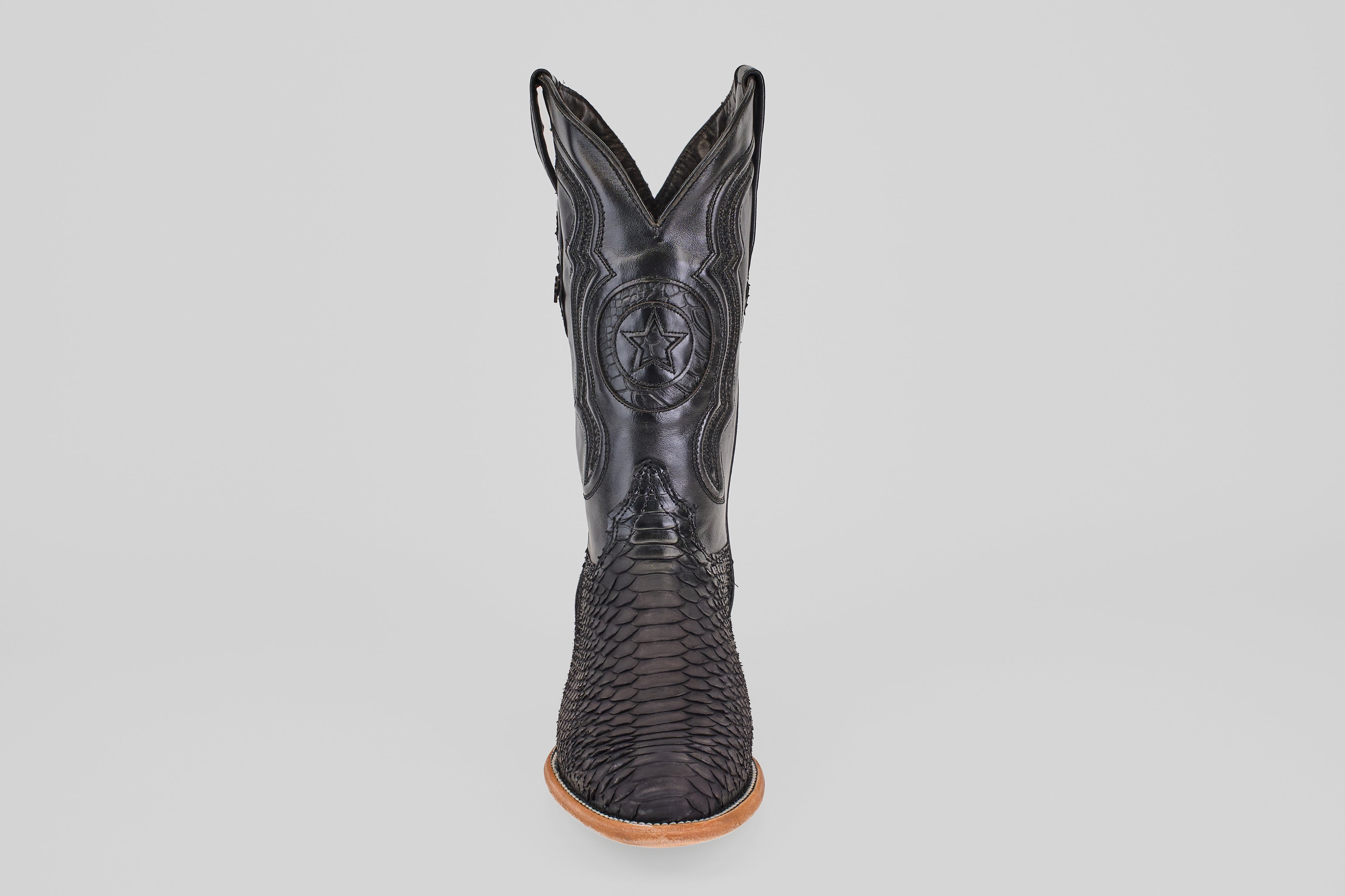 The Tanner – Matte Black – Roper Toe boot stands upright on a light brown sole, featuring a scaly texture and stitched star emblem on the shaft, showcasing western style against a clean white background.