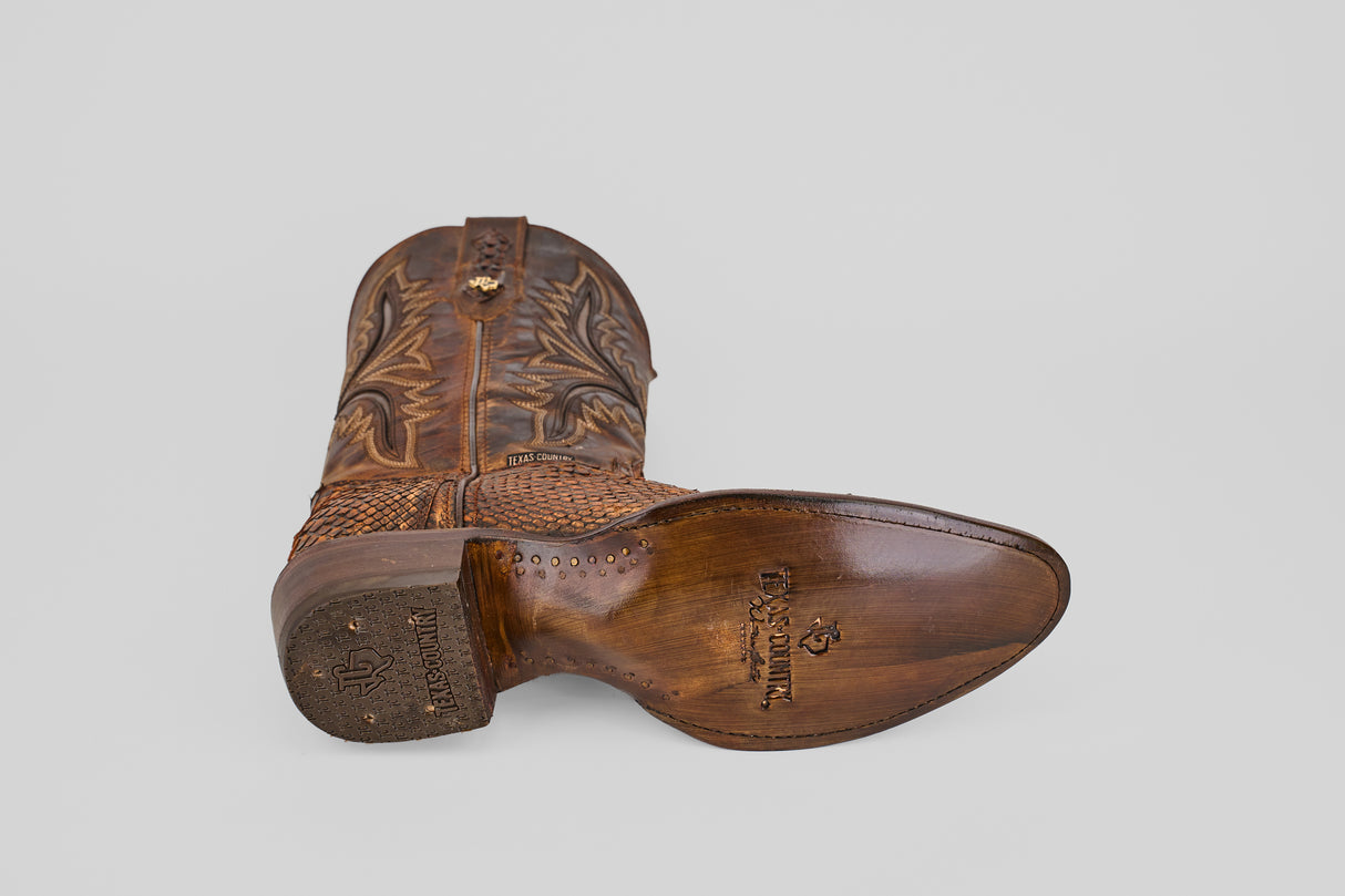 The Tanner – Chestnut – Round Toe boot rests on its side, displaying its detailed stitching and Lucchese-stamped sole. Set against a plain light gray background, it perfectly highlights classic western fashion style.