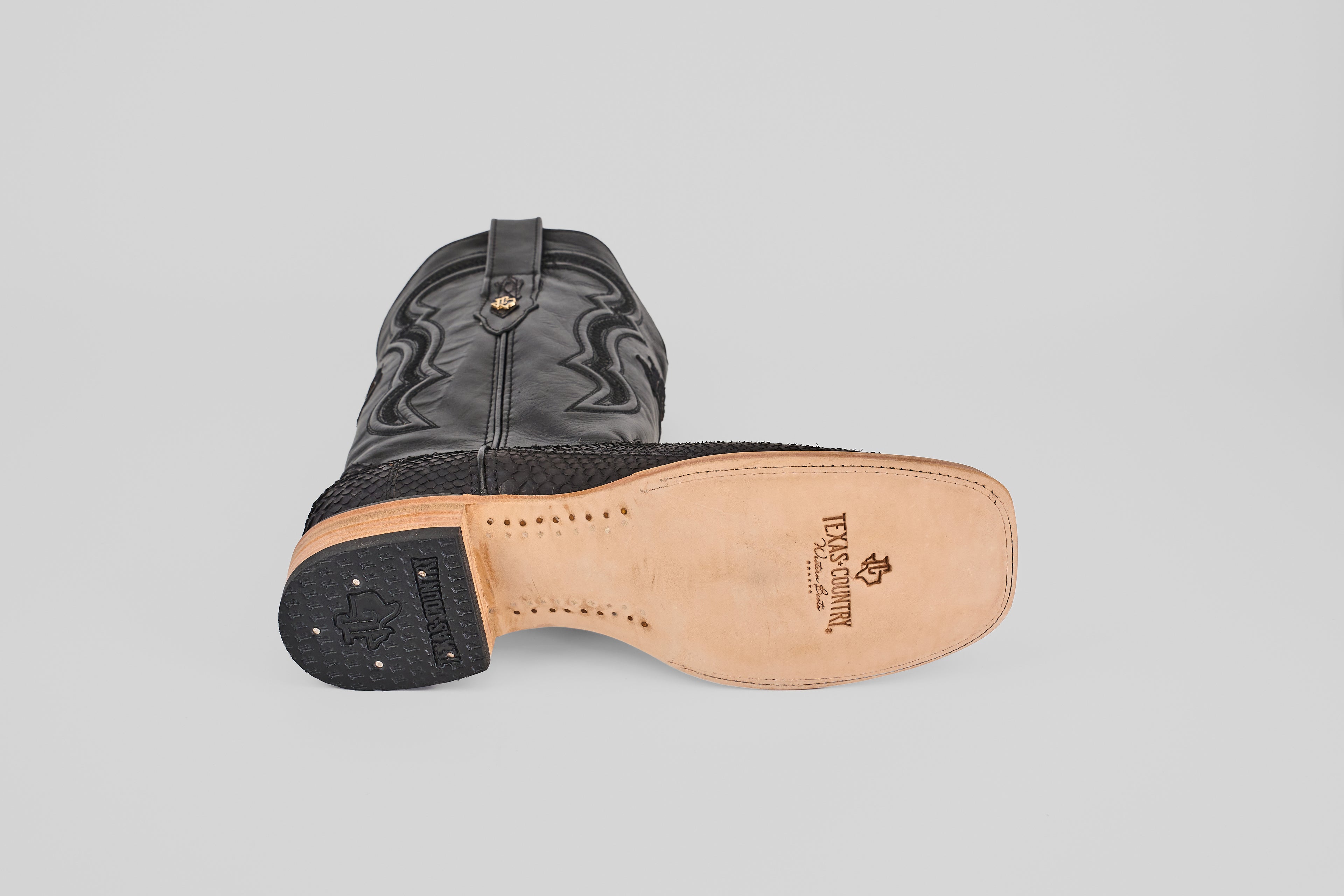 A Tanner - Matte Black - Square Toe cowboy boot lies on its side, displaying the textured matte black upper and the tan sole with brand and stitching details visible underneath.