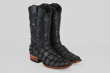 The Conor - Matte Black - Narrow Square Toe cowboy boots feature scale-like textured fish leather, detailed stitching, brown soles, and low wooden heels. They are shown on a light gray background.