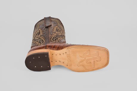 A single Hornback Brown - Embossed Leather - Square Toe (E427) boot is shown on its side, highlighting the detailed upper stitching and engraved logo with a patterned tan sole against a plain light gray background.