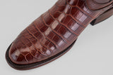 A close-up of the James Platimun - Dark Cogñac - Roper Toe (Diamond) shoe showcases its glossy brown crocodile pattern, detailed texture, and fine stitching along the sole against a light gray background.
