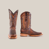 The Azkar Tan boots, featuring a Rodeo Toe and slip-resistant sole, are crafted from premium cowhide leather with intricate stitching. One boot is upright showing the side profile while the other stands front-facing, showcasing their Western elegance.