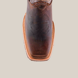 Close-up of the Azkar Tan boot with a Rodeo Toe, crafted from cowhide leather featuring intricate toe stitching and a light-colored welt. It has a squared design with a slip-resistant sole against a neutral background.