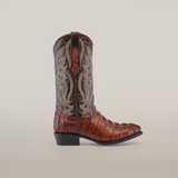 The Crocodile Tail Print Chocolate cowboy boot boasts a round toe with a crocodile tail design on the foot, ornate stitching on the shaft, and a decorative pull tab. Its angled heel stands out against the plain white backdrop.