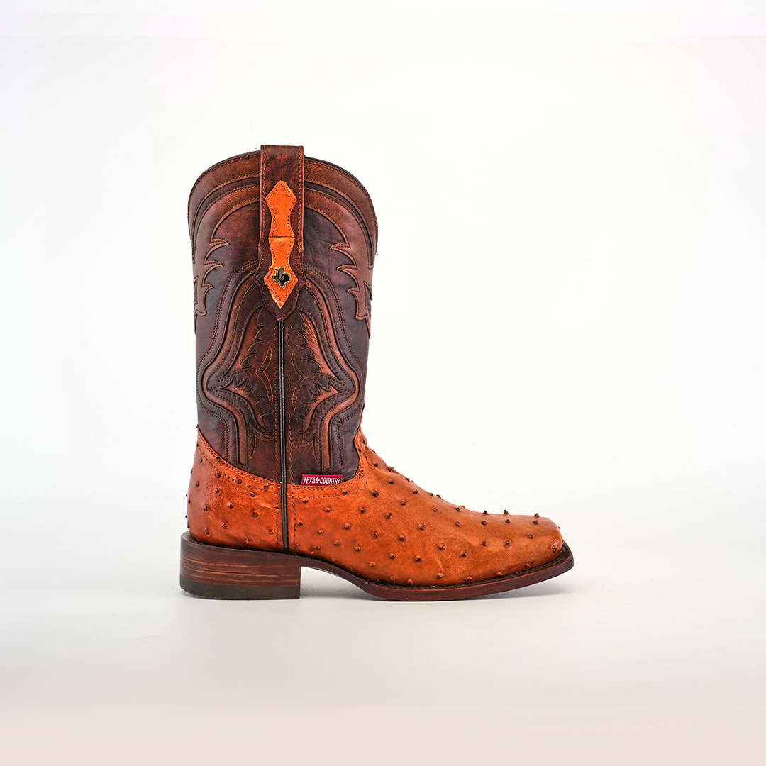 The Exotic Ostrich - Brandy boot features a light brown ostrich leather foot and a dark brown shaft with intricate stitching. Highlighting exceptional craftsmanship, it has a low heel and branded tag near the ankle, all beautifully set against a plain white background.