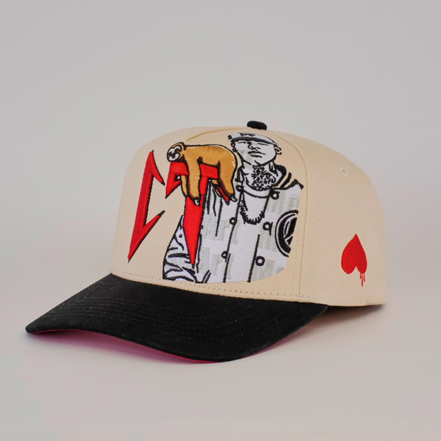 The De Rancho Cap CT Camel is a beige and black baseball cap featuring abstract red shapes, a monochrome illustration on the front, embroidered designs, and a red spade symbol on the side for a touch of Western heritage.