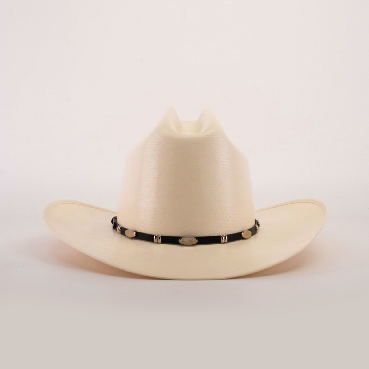 The 1000X Dos Carnales Straw Hat is a white cowboy hat with a wide brim and a black band adorned with silver conchos, crafted with exceptional quality. It stands out against a light background, proudly showcasing its superior craftsmanship.