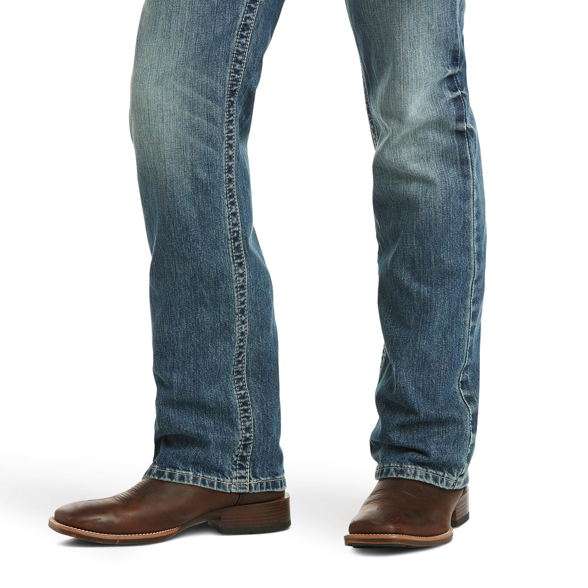 A pair of legs in M5 Slim Gambler Stackable Straight Leg Jeans with a slightly faded look and visible seams, paired with brown leather cowboy boots with low heels and rounded toes, displayed against a plain white background.