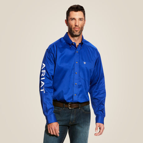 A man in a classic-fit Team Logo Twill Shirt Blue - 10017498 with ARIAT on the sleeve, blue jeans, and a brown belt, all premium cotton, stands against a plain background.