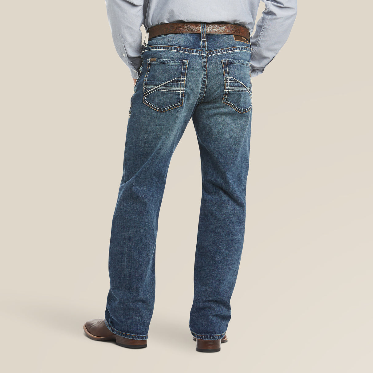 The individual is styled in Ariats M4 Low Rise Stretch Preston Boot Cut Jeans - Silverton - 10023455, with decorative back pocket stitching, a brown belt and shoes. The boot-cut jeans pair well with the light blue shirt against a plain light beige background.