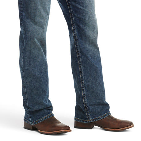 The image shows a person wearing Ariat M4 Low Rise Stretch Preston Boot Cut Jeans in blue denim (Silverton, 10023455) paired with brown leather cowboy boots. The slight fade and stretch denim highlight the pointed toe and low heel of the boots, focusing on the lower legs and feet.