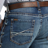 A close-up shows a person wearing Ariat M4 Low Rise Stretch Preston Boot Cut Jeans with detailed stitching and a branded label on the back pocket. A brown leather belt is threaded through the Silverton denims belt loops.