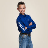 A young boy with a confident smile and short brown hair stands against a plain, light-colored background, wearing an Ariat Boys Blue Team Logo Long Sleeve Western Shirt - 10030164 made from durable cotton-blend twill. With Ariat on the sleeve, he crosses his arms over the blue button-up.