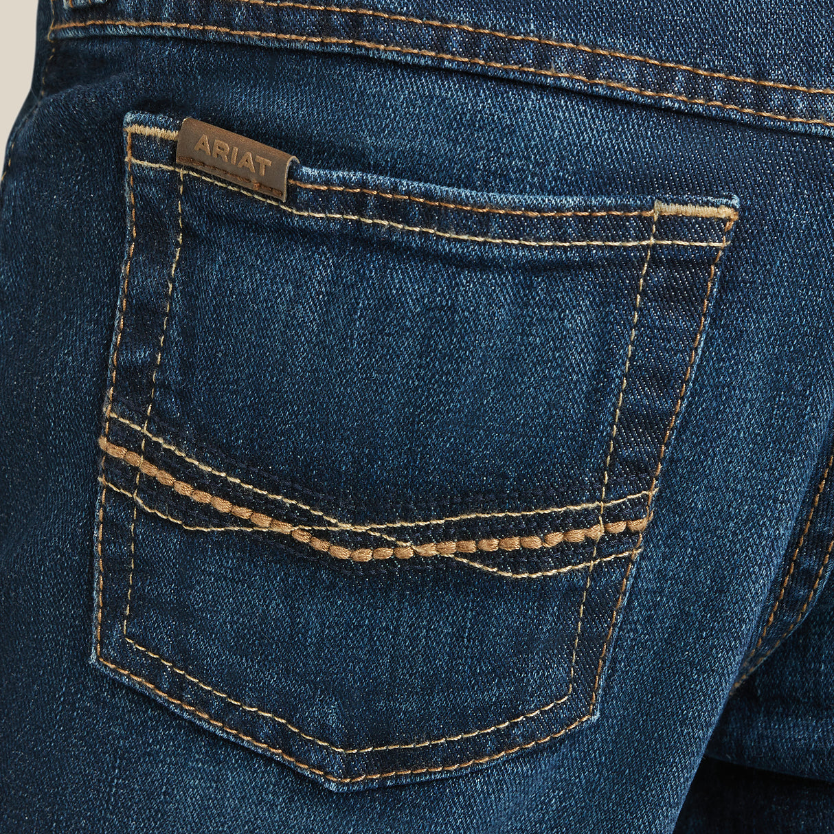 A close-up of the back pocket of Ariat Boys B4 Legacy Dark Wash Relaxed Fit Boot Cut Jeans, featuring detailed stitching with a small brown leather patch reading Ariat. The premium denim has a worn texture for a casual look.