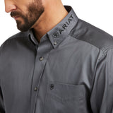 A man wearing the Team Logo Twill Classic Fit Shirt Gray - 10037479, featuring Ariat and a logo embroidered on the collar and pocket with smooth fabric, is looking down slightly. The shirt also has a front pocket.