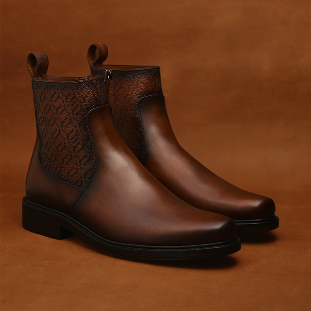 Introducing the Honey Laser - Round Toe, ankle boots crafted from polished brown leather featuring intricate laser-cut patterns on the sides and handy pull tabs. These boots showcase their premium materials against a warm, matching brown backdrop.