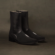 The Black Stingray Zipper - Round Toe boots feature sleek black leather with a side zipper and textured front panel, complemented by a low heel and set against a dark, mottled background.