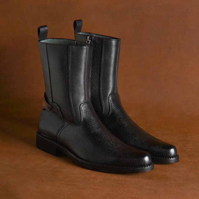 The Black Deer Zipper - Round Toe boots from the CUADRA premium collection are elegantly displayed on a brown surface. These black leather boots feature a sleek, smooth finish, adjustable side straps, and a slight heel for a polished and sophisticated look.