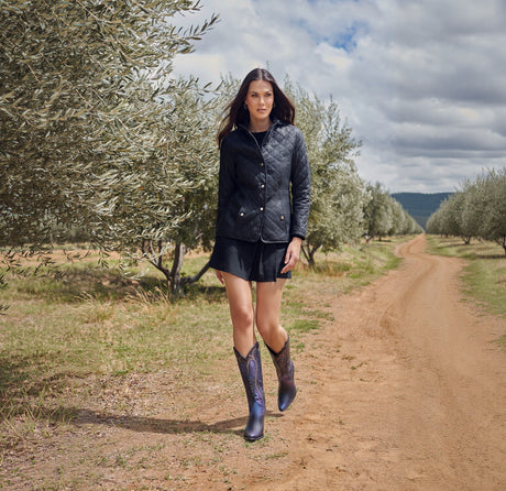 A woman in a quilted jacket with floral embroidery and Black Overlay & Embroidery & Studs - Snip Toe cowboy boots strolls along a tree-lined dirt path under a cloudy sky.