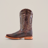 The Cheyenne Chocolate boot, made from premium cowhide, features intricate stitching on its dark brown shaft. Its rodeo toe and stacked wooden heel add rustic elegance, while the slip-resistant sole balances style with practicality against a plain white backdrop.