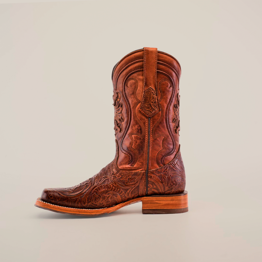 A single handcrafted cowboy boot, the Handtooled Leave Brown features a slip-resistant sole, rodeo toe, and intricate stitching on premium brown cowhide leather with a medium wooden heel. Set against a light beige background, it includes pull straps for easy wear.